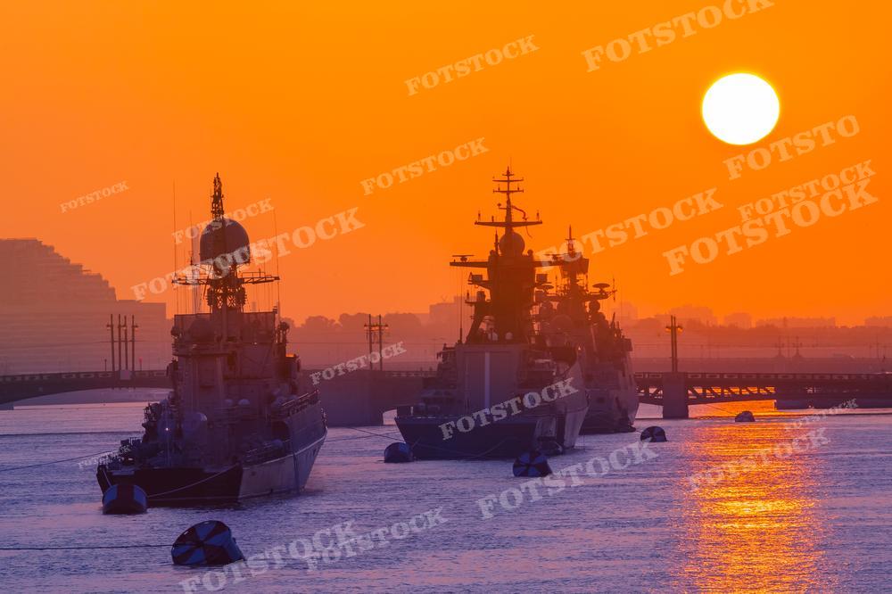 На фотографии изображены военные корабли на фоне заката, солнце садится, окрашивая небо в оранжевые тона. За кораблями видны очертания городских сооружений и моста. Морской пейзаж в оранжевых тонах может использоваться для иллюстрации статей о военно-морском флоте, в качестве обложек книг по морской тематике, в фотоискусстве и дизайне, а также для создания атмосферных картин и постеров.
